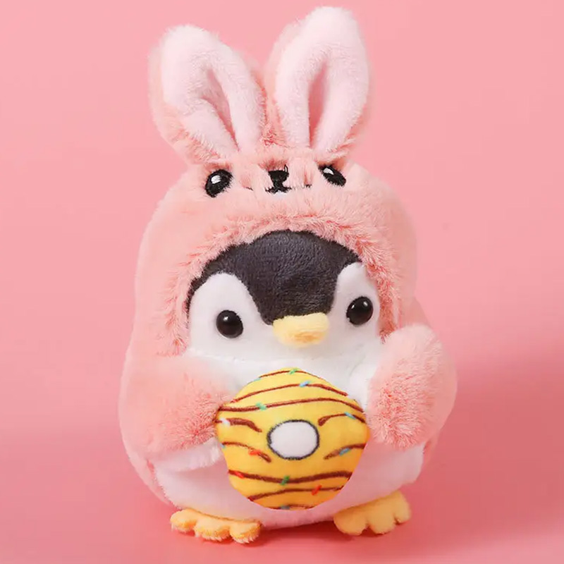 Kleiner Plüschpinguin, der ein rosafarbenes Hasenkostüm trägt und einen orangefarbenen Donut in den Händen hält.