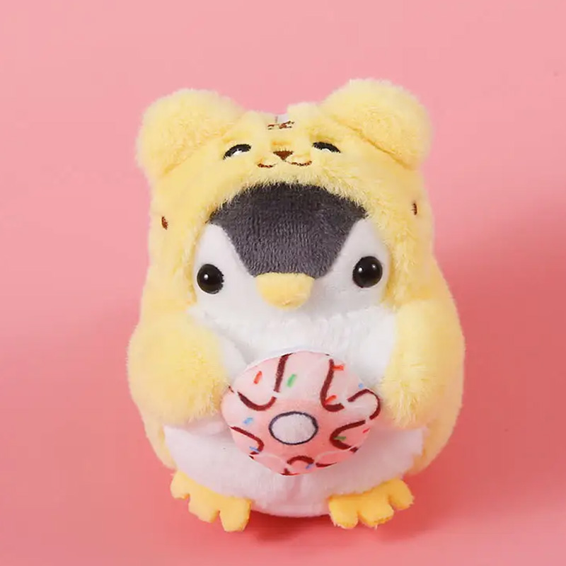 Schlüsselanhänger Pinguin im Pyjama 10cm Kleiner Plüschpinguin, der ein gelbes Bärenkostüm trägt und einen rosafarbenen Donut in den Händen hält.