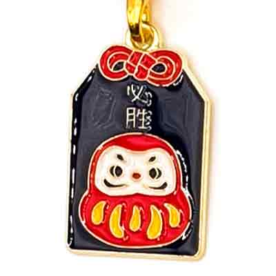 Goldener Anhänger in Form eines japanischen Geschenkanhängers. Darauf ist eine rot-gelbe Puppe auf schwarzem Hintergrund abgebildet.